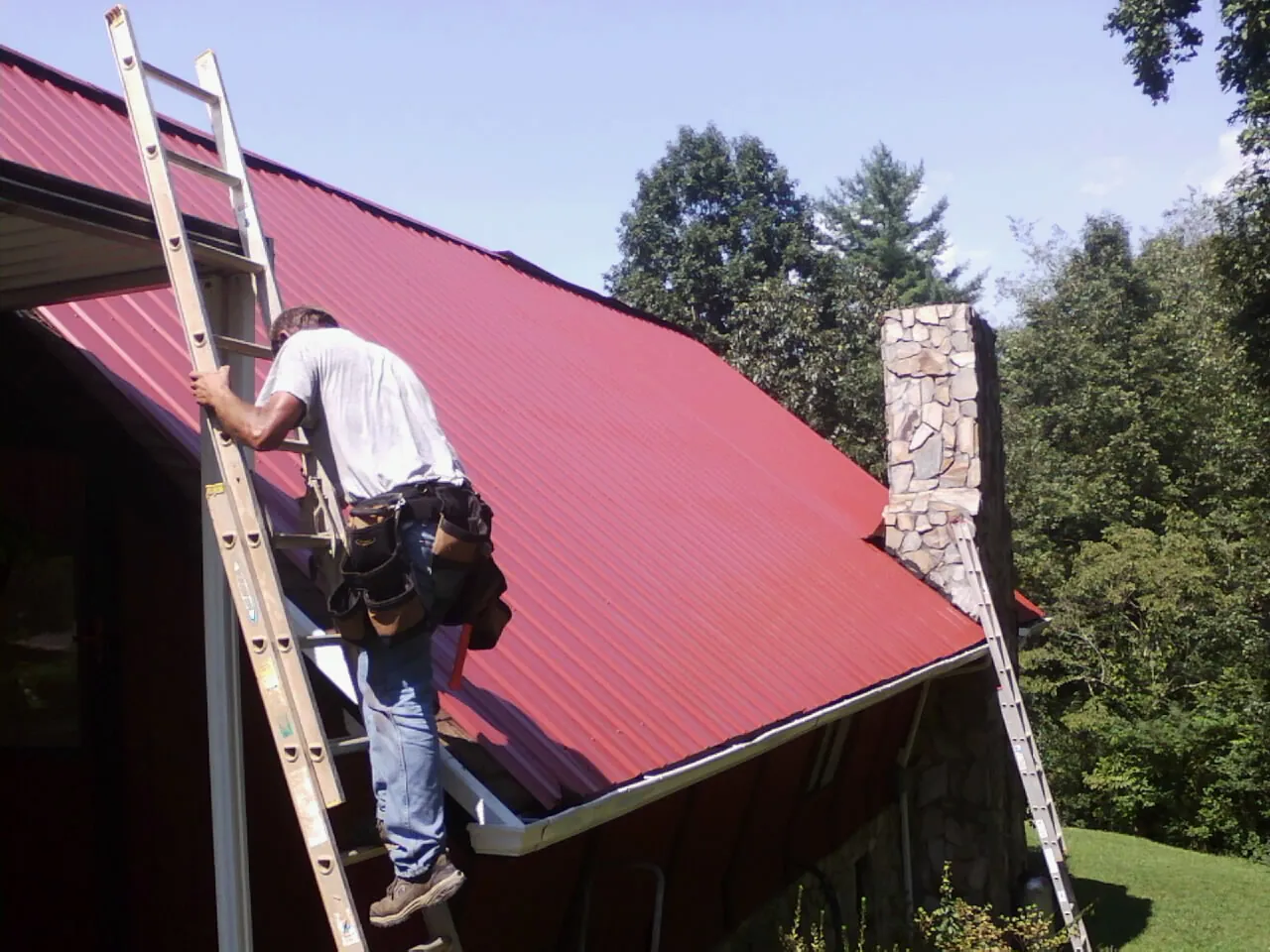 Roofer on a ladder beside a red metal roof and stone chimney — Soffit & Fascia Repair by Roofing Brothers in Ipava